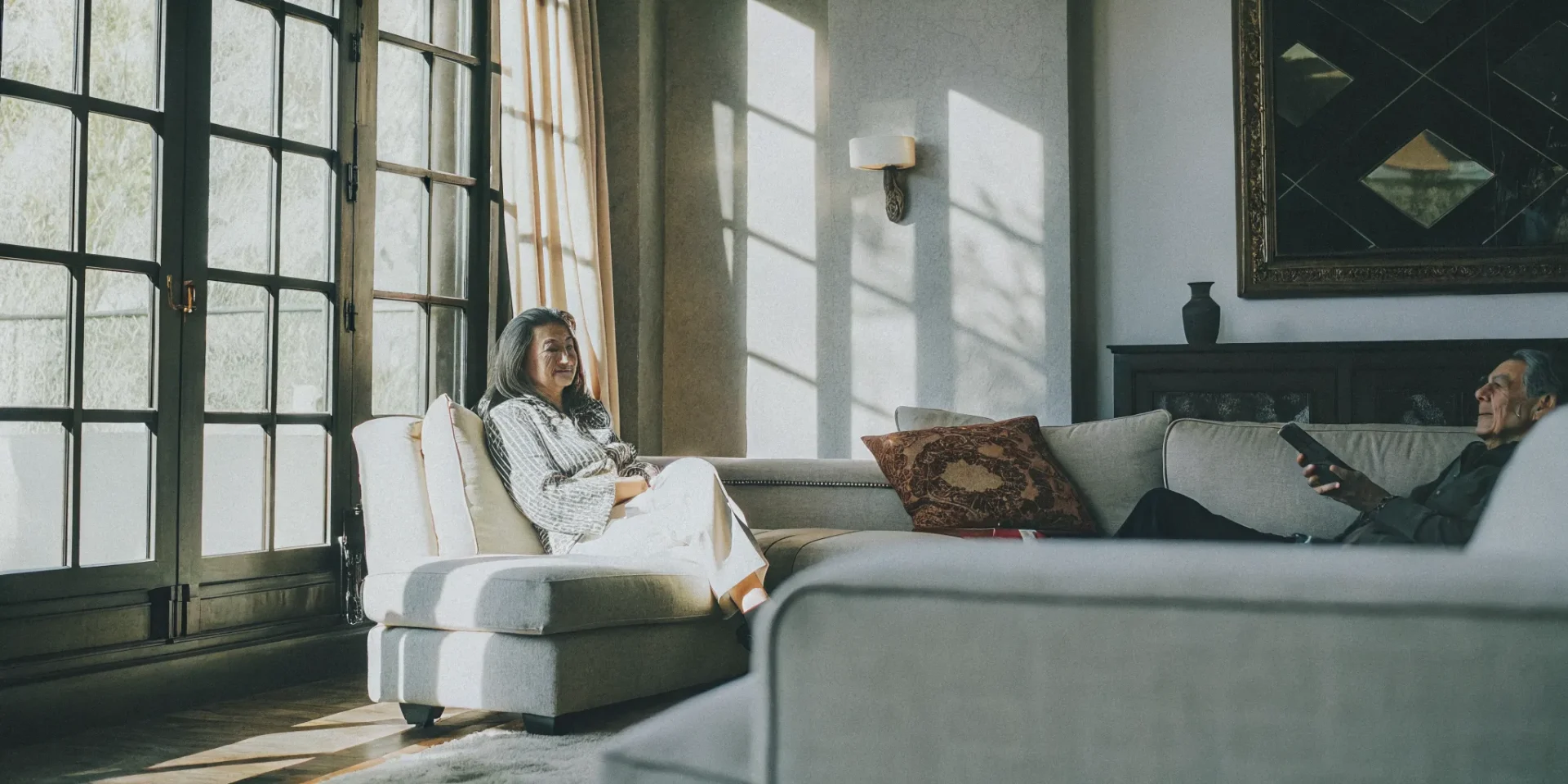 Elderly woman sitting with a companion in a bright, elegant living room with natural light streaming through tall windows